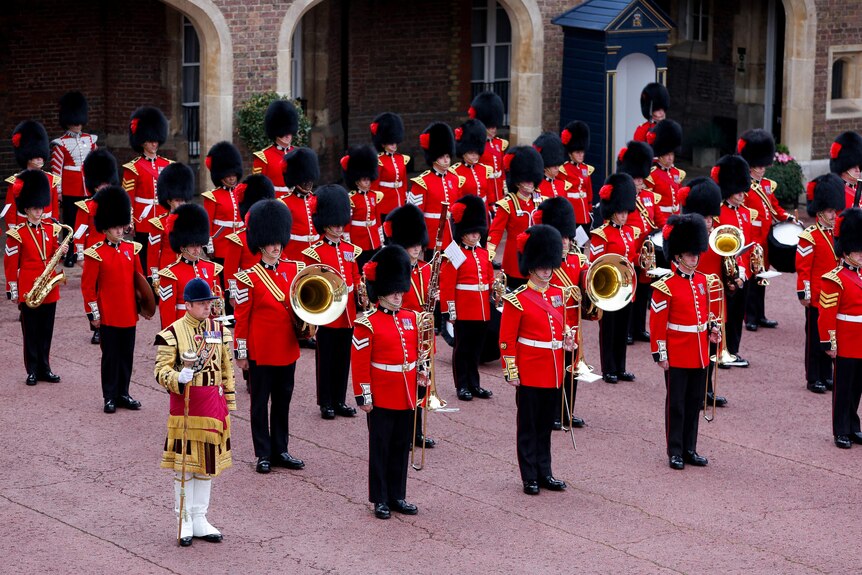 Buckingham Palace guards in red uniform stand in rows with various marching band instruments. 