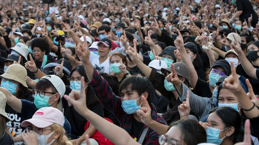 Thousands of protesters, many wearing disposable face masks, raise their right hands and point index fingers up.