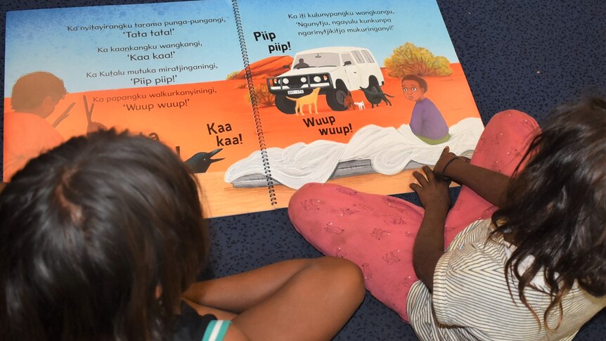 Looking over the shoulders of two Aboriginal children sitting next to each other reading a book in language.