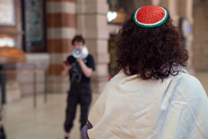 Back of person's head, which has a watermelon-coloured kippah on top of hair. In the background, a person uses a megaphone.