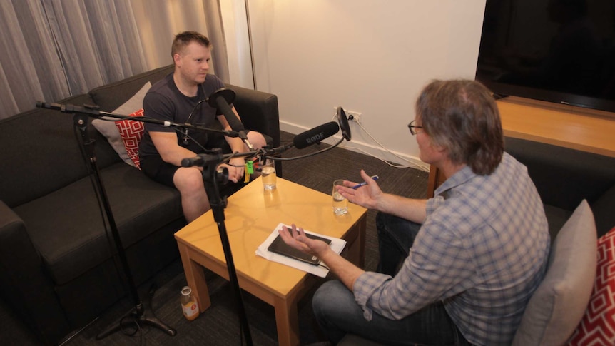 Chris Hill sitting on a couch being recorded by Angus Grigg (back to camera)