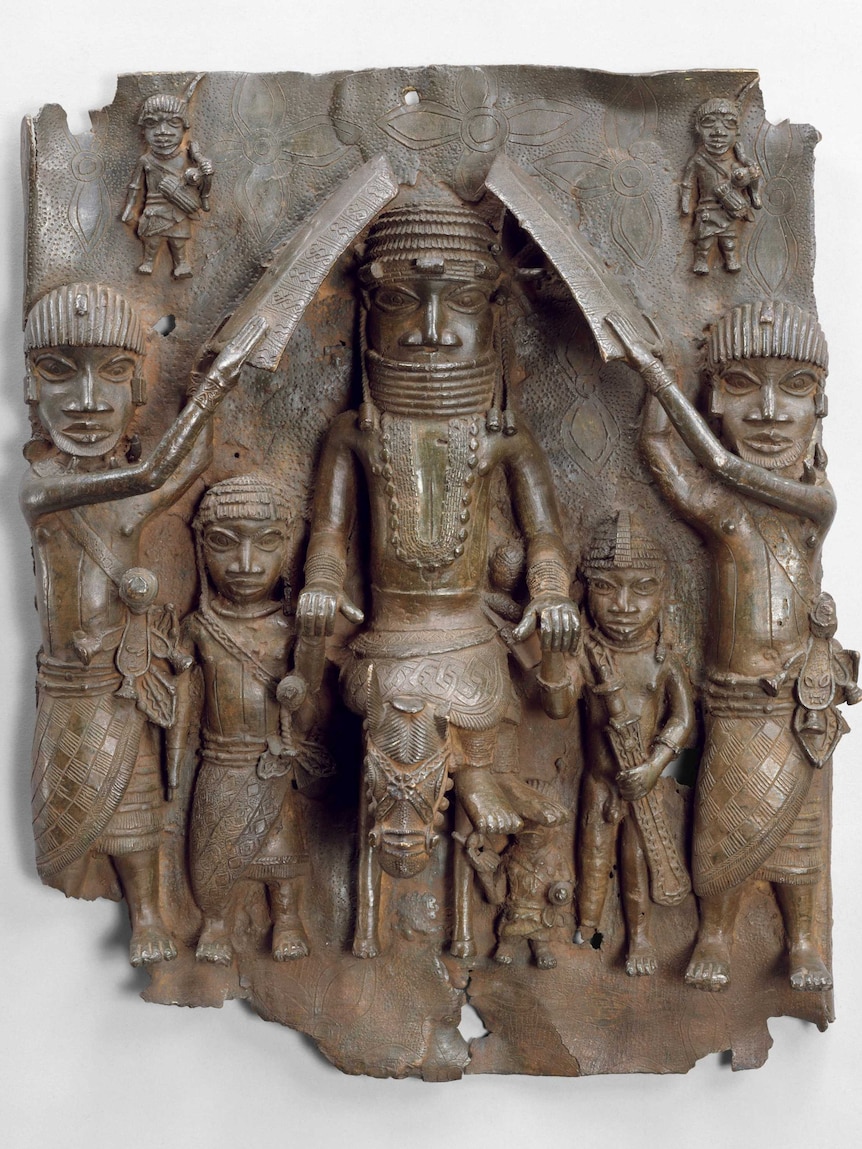 A brass sculpture on a plaque of a Benin king on a horse, attendants fan the king and walk