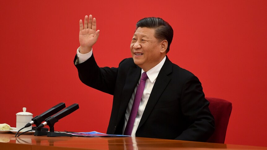 Xi Jinping waves to someone off camera as he sits in a suit in front of a red wall.
