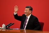 Xi Jinping waves to someone off camera as he sits in a suit in front of a red wall.