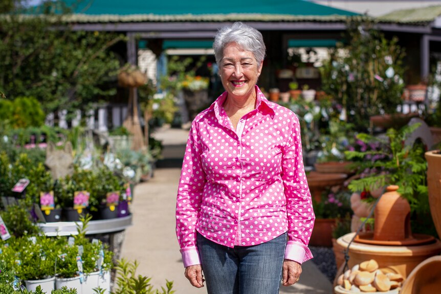 A smiling woman in a pink top stands in a plant nursery