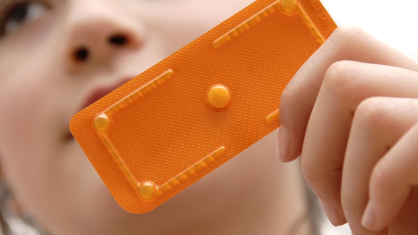 A woman holding a single pill in a bright orange packet.