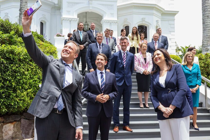 A man takes a selfie with a group of people standing in front of a white building.