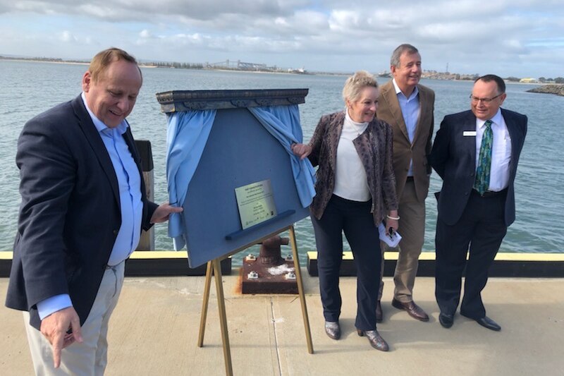 One woman and three men in suits stand smiling next to a board with a plaque on it on a jetty