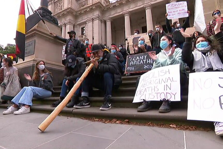 A crowd of people, mainly wearing masks and holding protest signs, sits on the steps of Victorian Parliament.