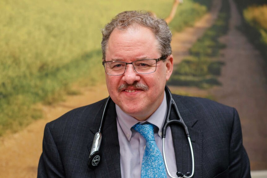 Head and shoulders shot of a doctor wearing a suit, stethoscope, glasses and a moustache.