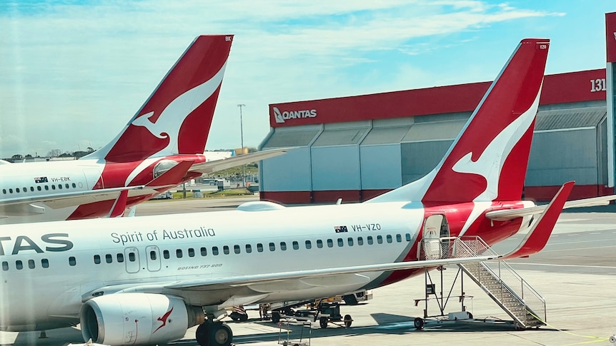 Two Qantas planes parked on a tarmac at an airport with a Qantas-branded shed in the background.