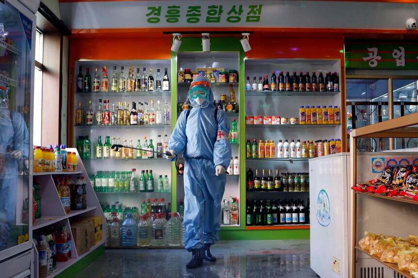 A person dressed in full personal protective equipment disinfects an aisle in a small grocery store.