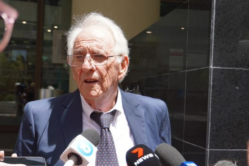 An old man in a suit and tie speaks to reporters. 