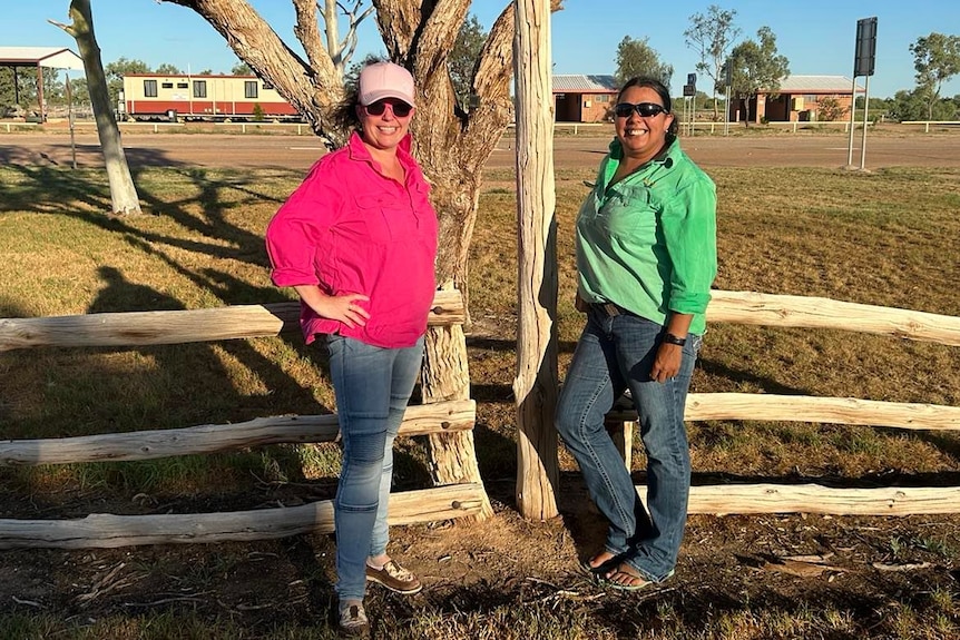 a woman in a pink shirt stands next to a woman in a green shirt in a dry looking outback street