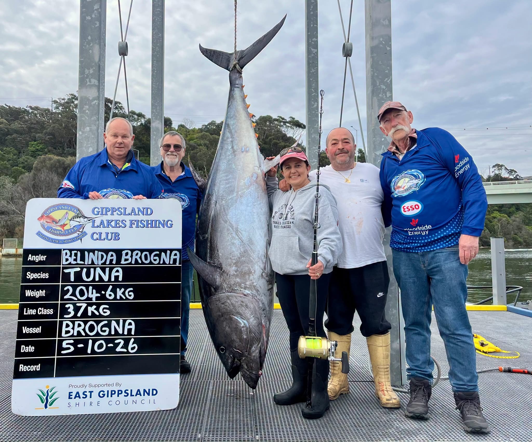 A group of five people smiling next to a giant tuna fish, standing on a jetty, water in background