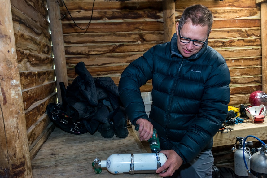 A man in a puffer jacket holding a scuba tank in a log cabin.