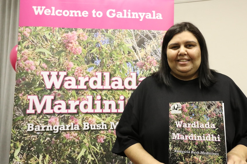 A smiling Indigenous woman in front of a large banner bearing the cover of a book about bush medicine.
