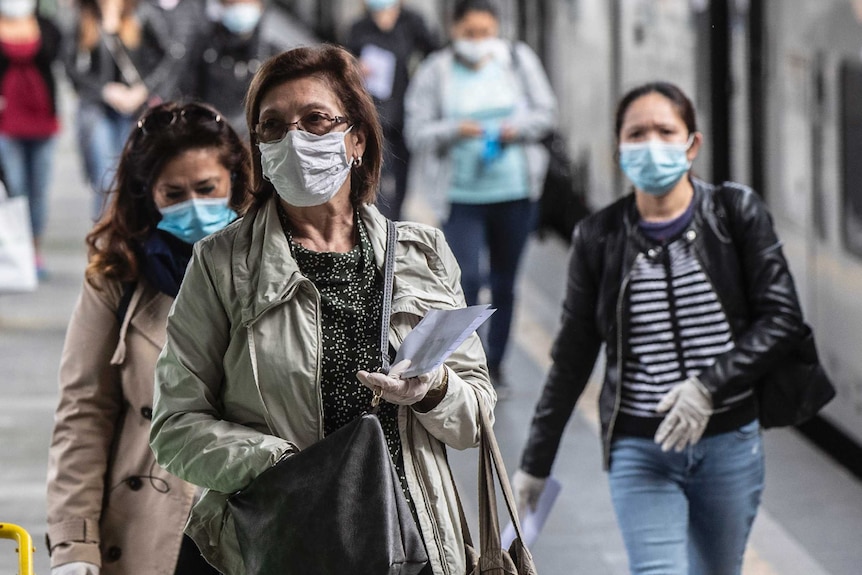 Women wearing face masks and rubber gloves walk past a train on a platform.