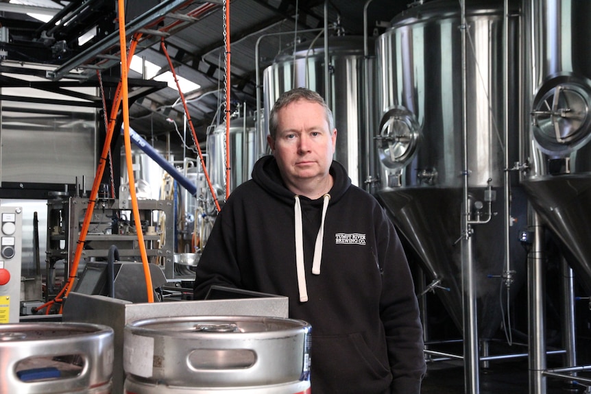 A middle-aged man in a black hoodie, standing behind a keg of beer in a brewery.