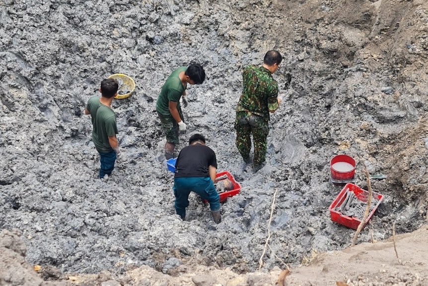 Five men stand in the grave pit with red buckets, trawling through the rubble to find artefacts and bodies.