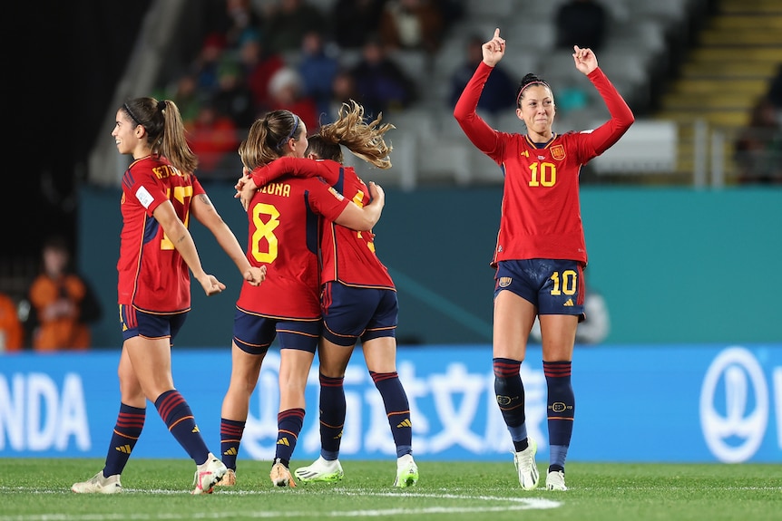 A Spanish striker points her fingers in the air in celebration after having a goal confirmed, as teammates embrace behind her.