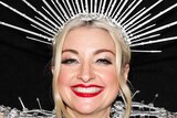 Kate Miller-Heidke, wearing an ornate silver head dress and clothing, poses for a photo smiling wide with her arms up in the air