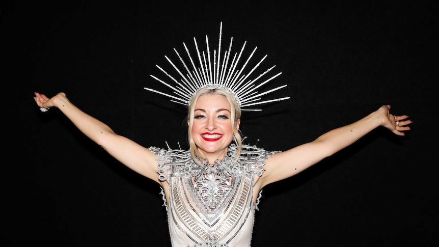 Kate Miller-Heidke, wearing an ornate silver head dress and clothing, poses for a photo smiling wide with her arms up in the air