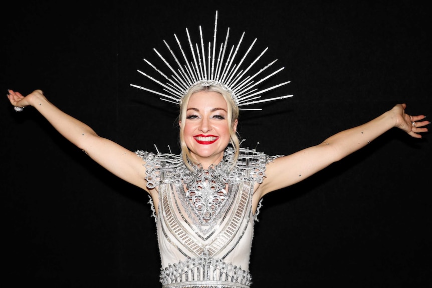 Kate Miller-Heidke, wearing an ornate silver head dress and clothing, poses for a photo smiling wide with her arms up in the air