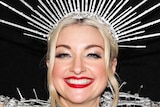 Kate Miller-Heidke, wearing an ornate silver head dress and clothing, poses for a photo smiling wide with her arms up in the air