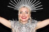 Kate Miller-Heidke, wearing an ornate silver head dress and clothing, poses for a photo smiling wide with her arms up in the air