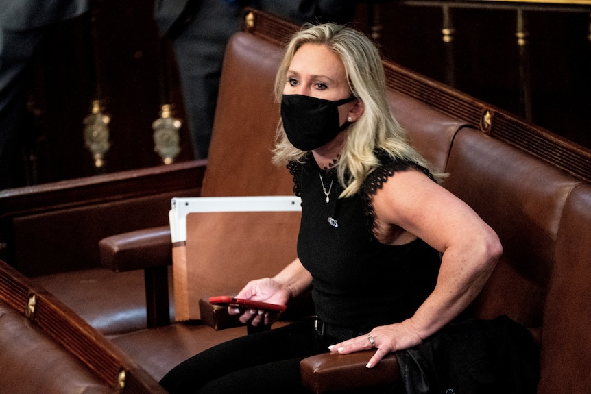 Marjorie Taylor Greene holds her phone while sitting in a brown leather seat. She's wearing a black dress and black face mask