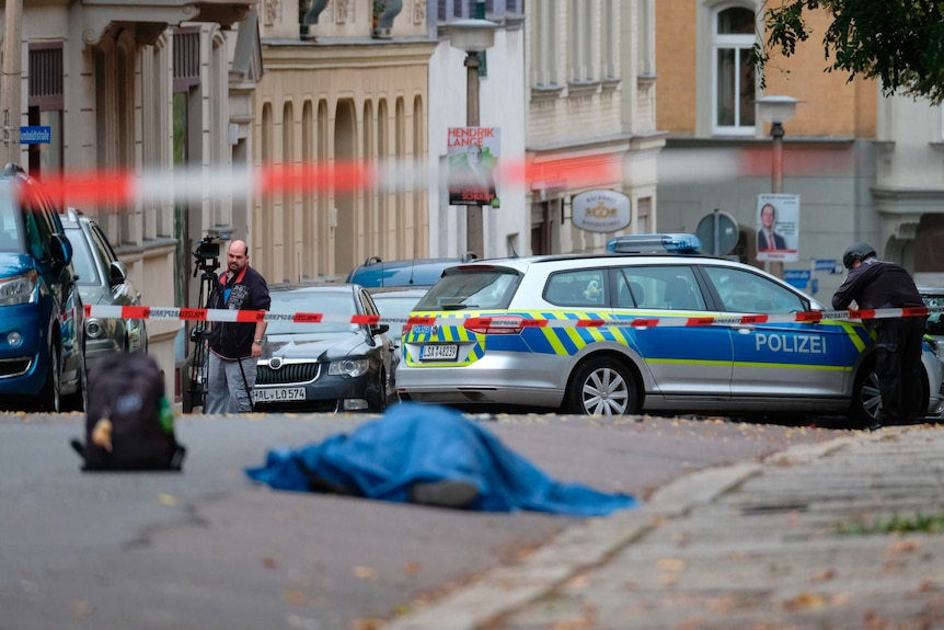 A dead body lies under a blue tarp, with the street taped off by police