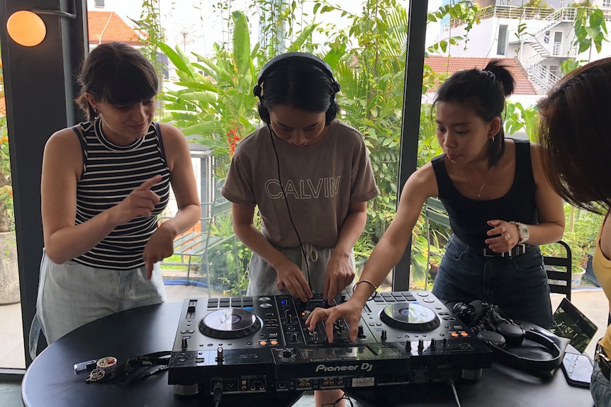 Three women huddle around a DJ deck, one dancing along.