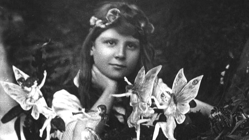 A black and white portrait of a young girl in Edwardian dress and flowers in her hair surrounded by small winged figures.