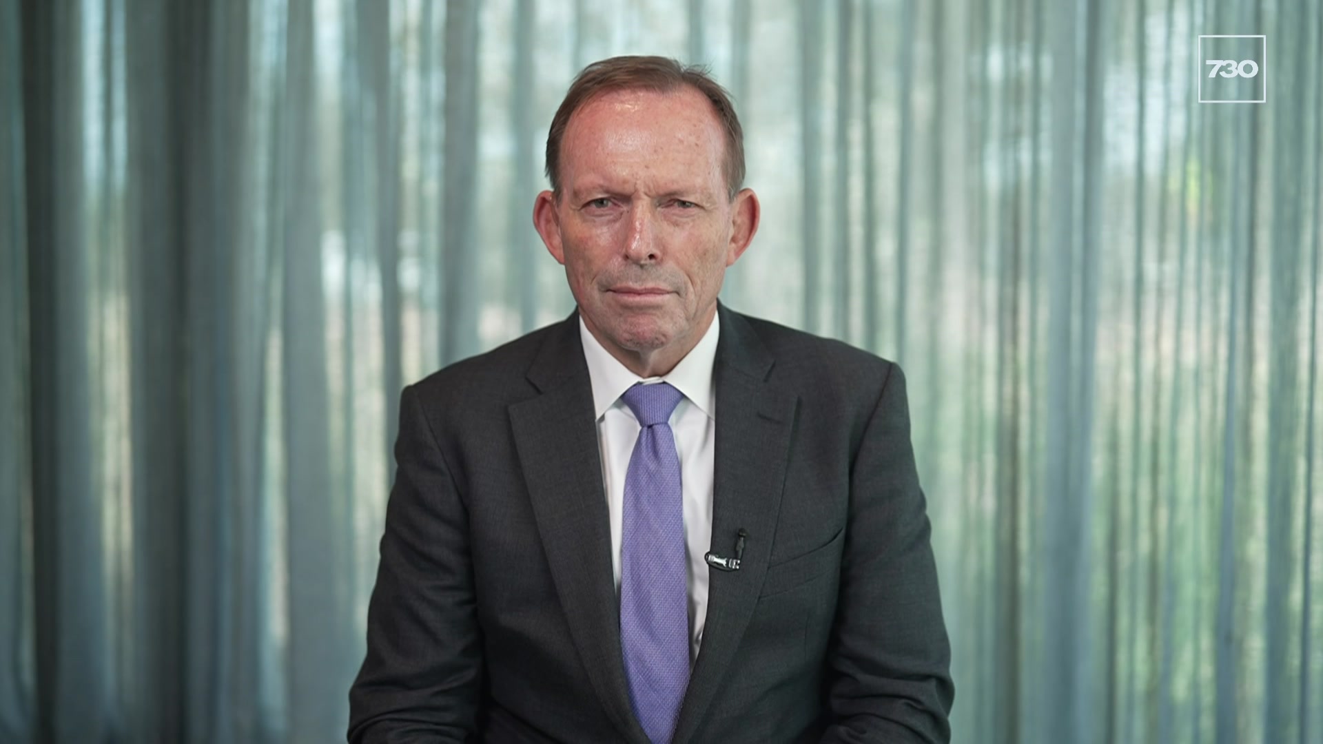 Tony Abbott wearing a suit with a purple tie sits in front of a window with sheer curtains. 