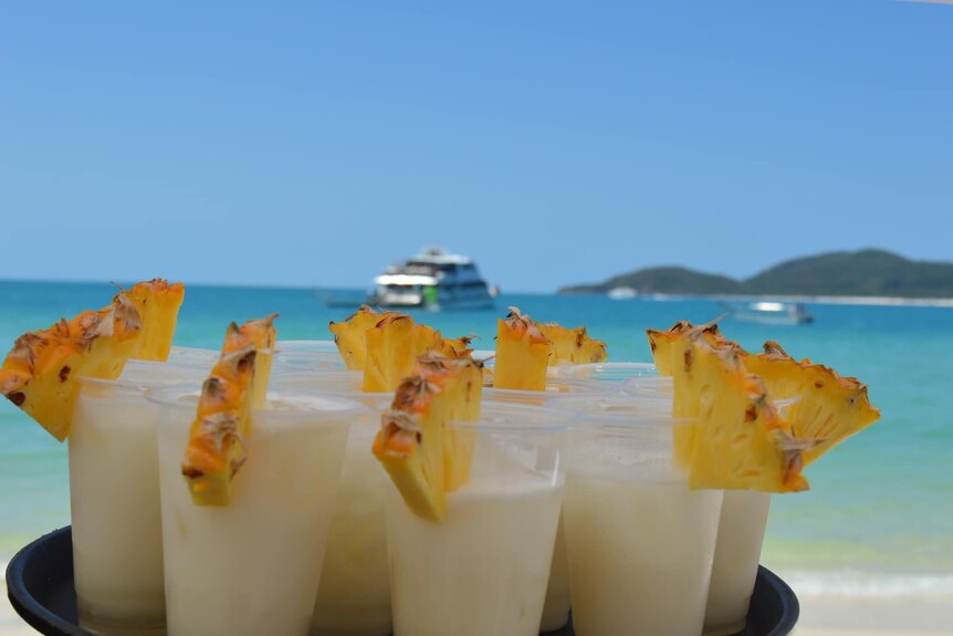 A plate of drinks with pineapple on the side of the glass are held in front of a blue ocean and sky. 