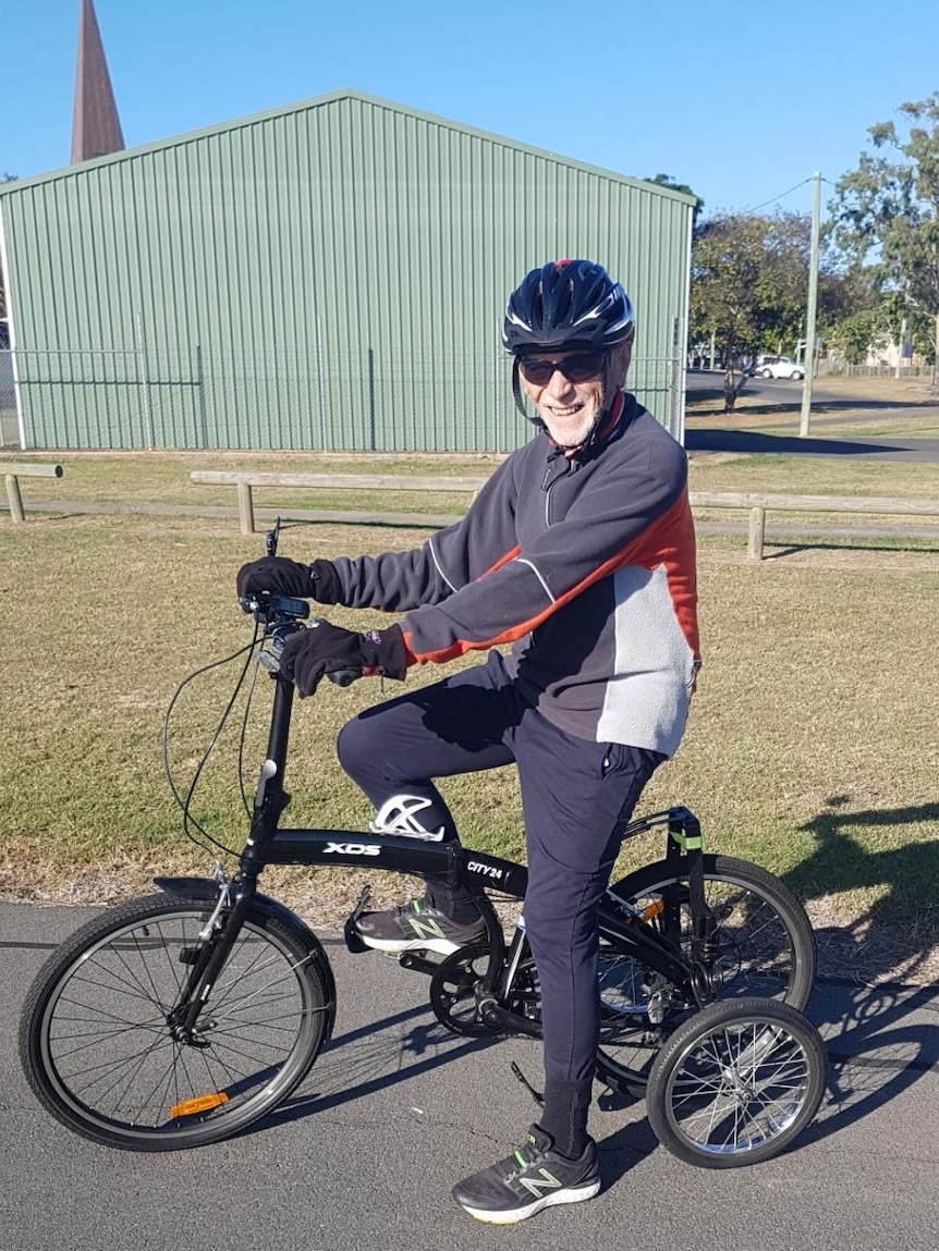 A man in a tracksuit, helmet and sunglasses astride a bicycle with large training-style wheels. He is smiling.