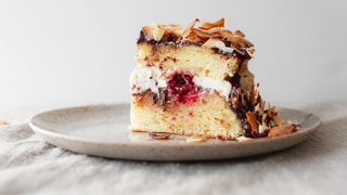 Slice of lamington cake with toasted coconut, cream, jam, fresh raspberries and chocolate ganache on a plate.