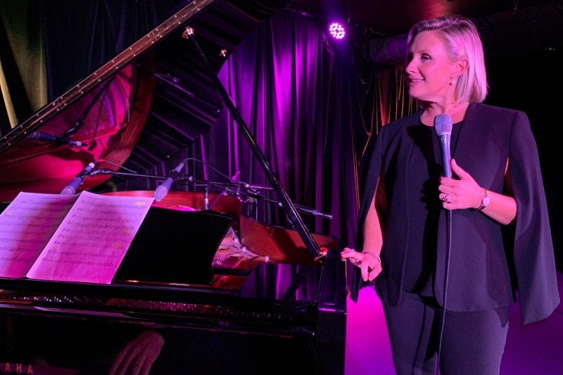 Woman stands to the side of piano with microphone