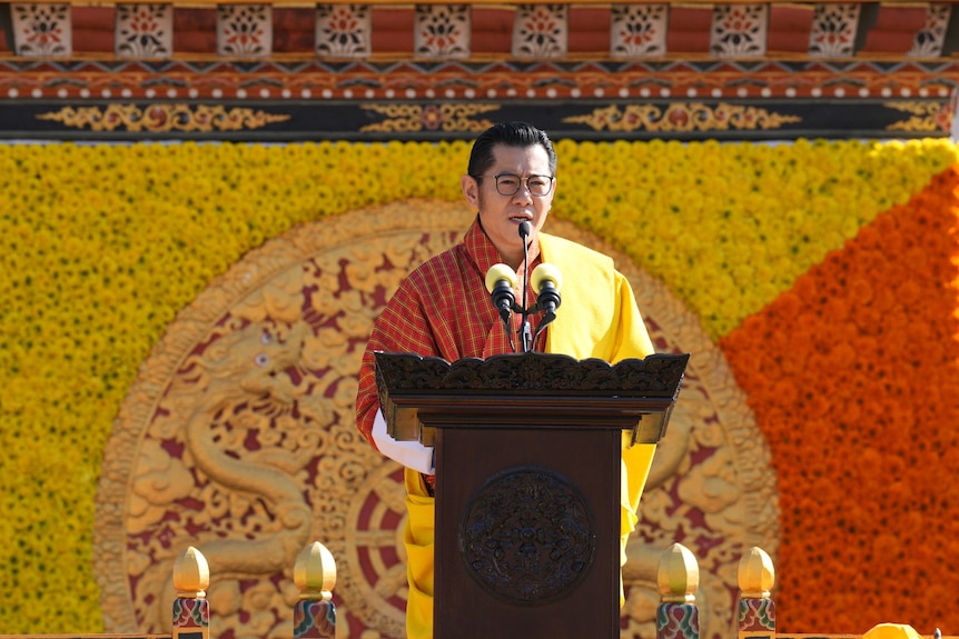  King Jigme Khesar stands behind a lectern wearing traditional Bhutanese clothing with a yellow and orange display behind him. 