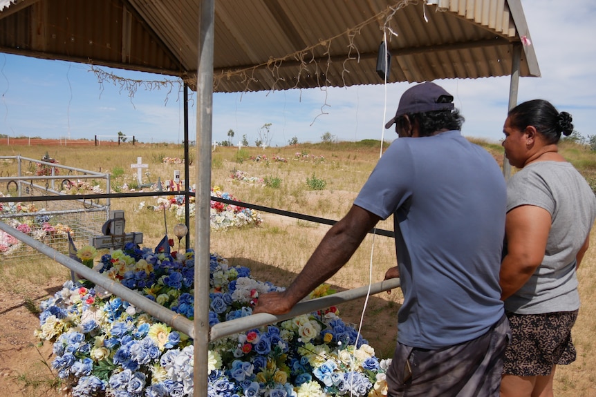 A man and woman standing infront of a grave covered in blue and yellow flowers.