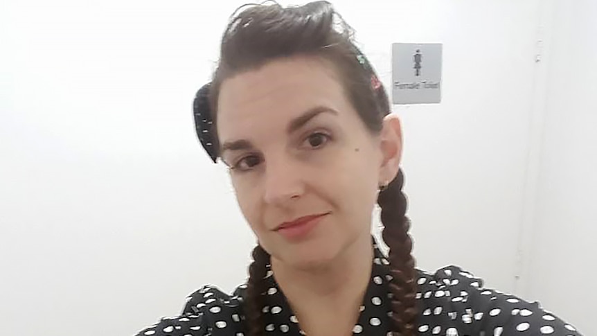 A woman with dark hair and a polka dot top takes a selfie shot with a public toilet door behind her.