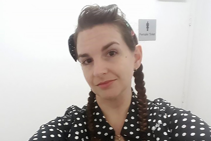 A woman with dark hair and a polka dot top takes a selfie shot with a public toilet door behind her.