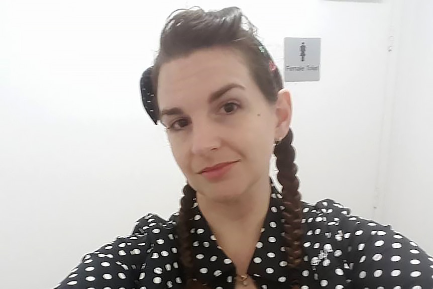 A woman with dark hair and a polka dot top takes a selfie shot with a public toilet door behind her.