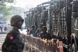 Soldiers stand next to military vehicles as people gather to protest.