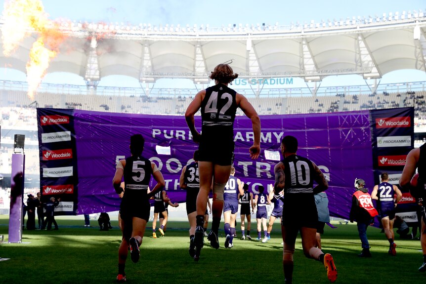 Fremantle Dockers players seen from behind running onto Perth Stadium for an AFL game with a purple banner ahead of them.