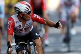 A cyclist in red white and black uniforms holds up his left index finger in celebration of a stage win.