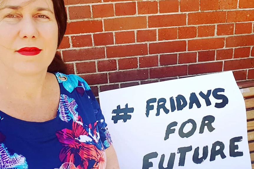 A woman smiles in front of a brick wall holding a sign that says hashtag Fridays for future