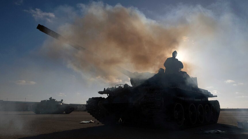 From a low angle, you look up at a silhouette of a tank with a man standing on top of it with artillery smoke engulfing the area
