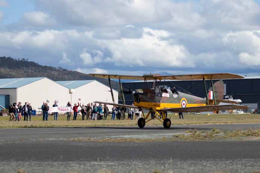 A crowd stands nearby a Tiger Moth plane on an airfield. 
